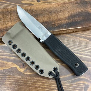 May include: A stainless steel knife with a black textured handle and a tan sheath. The knife has a pointed blade and is laying next to its sheath. The sheath has a series of eyelets along the edge and a black cord.