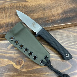 Pode incluir: Uma pequena faca com lâmina prateada e cabo preto texturizado, acompanhada por uma bainha verde com ilhós e um cordão preto. A lâmina tem a inscrição "PENDLETON MINI HUNTER".