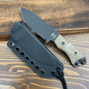 Könnte beinhalten: Ein schwarzes Messer mit feststehender Klinge, einem beigefarbenen Griff und einer schwarzen Scheide. Auf der Klinge steht "BK&T KA-BAR". Die Scheide ist schwarz mit Ösen und einer schwarzen Kordel.