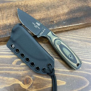 Può includere: Un coltello Izula-II nero con lama nera e manico verde e marrone, appoggiato su una superficie di legno. Il coltello è accompagnato da un fodero nero con occhielli e un cordino. La lama riporta la scritta "ESEE KNIVES USA" e "IZULA-II".