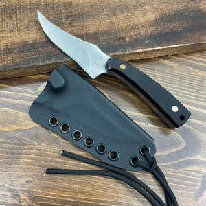 May include: A stainless steel knife with a curved blade and black handle rests beside its black sheath. The knife has a small, pointed blade and a black handle with a brass rivet. The sheath has eyelets for a cord.