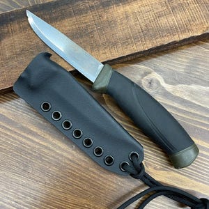 May include: A survival knife with a silver blade and a black handle, accompanied by a black sheath. The knife is designed for outdoor use and survival situations. The sheath has multiple eyelets and a black cord.