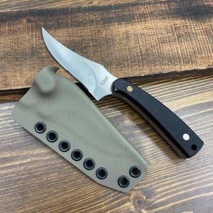 Può includere: Un coltello a lama fissa con una lama in acciaio color argento contrassegnata 1520T e un manico nero testurizzato fissato con due rivetti in metallo. Accanto al coltello c'è un fodero sagomato color marrone chiaro con sei occhielli neri sul bordo.