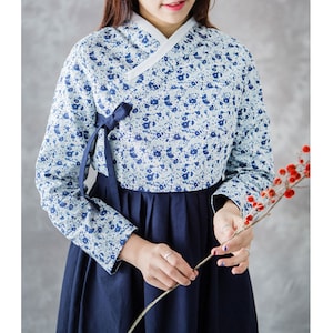 Op de afbeelding: Een traditionele Koreaanse hanbok-jurk. De top is lichtblauw met een bloemenpatroon en witte bies, gecombineerd met een donkerblauwe rok. Een donkerblauw lint is op de borst geknoopt. De mouwen zijn lang en de jurk is gemaakt van een zachte stof.