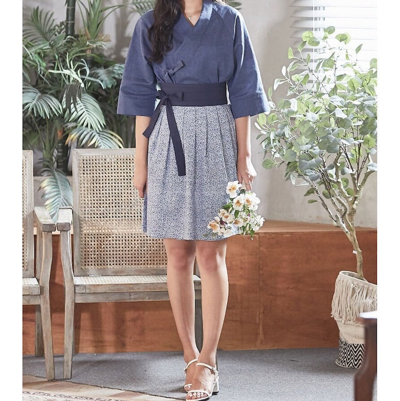 May include: A navy blue top with a V-neck and three-quarter sleeves, paired with a blue and white floral print skirt. A dark blue sash wraps around the waist. The outfit is completed with white sandals and a bouquet of white flowers.
