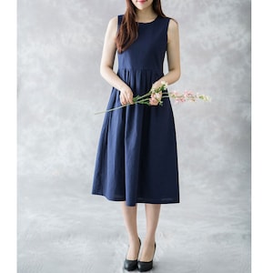 Op de afbeelding: Een mouwloze, marineblauwe jurk met een geplooide taille en een uitlopende rok. De jurk is knielang en wordt gedragen met zwarte hakken. De persoon houdt een klein boeket roze bloemen vast.