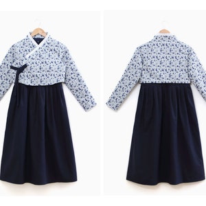 Op de afbeelding: Een traditionele Koreaanse Hanbok-jurk. De top is lichtblauw met een bloemenpatroon en lange mouwen. De rok is effen marineblauw. De jurk wordt gepresenteerd op een houten hanger tegen een witte achtergrond.