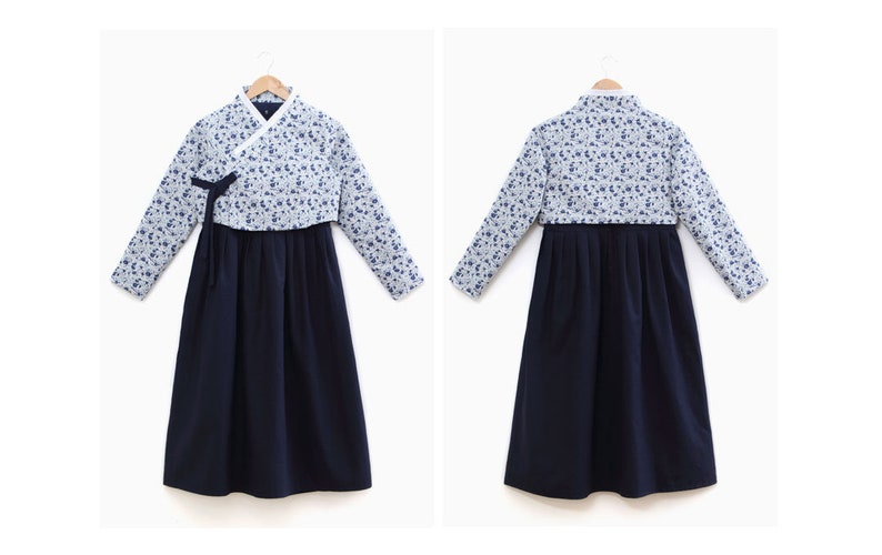 Op de afbeelding: Een traditionele Koreaanse Hanbok-jurk. De top is lichtblauw met een bloemenpatroon en lange mouwen. De rok is effen marineblauw. De jurk wordt gepresenteerd op een houten hanger tegen een witte achtergrond.