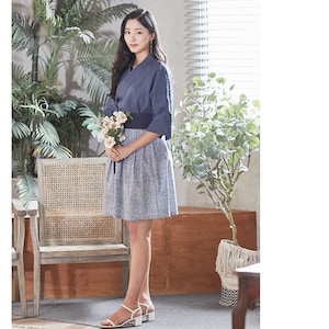 May include: A woman wearing a navy blue wrap top with three-quarter sleeves, a matching wide belt, and a blue floral print skirt. She is holding a bouquet of white flowers and wearing white sandals with block heels. The outfit is suitable for a casual or semi-formal occasion.