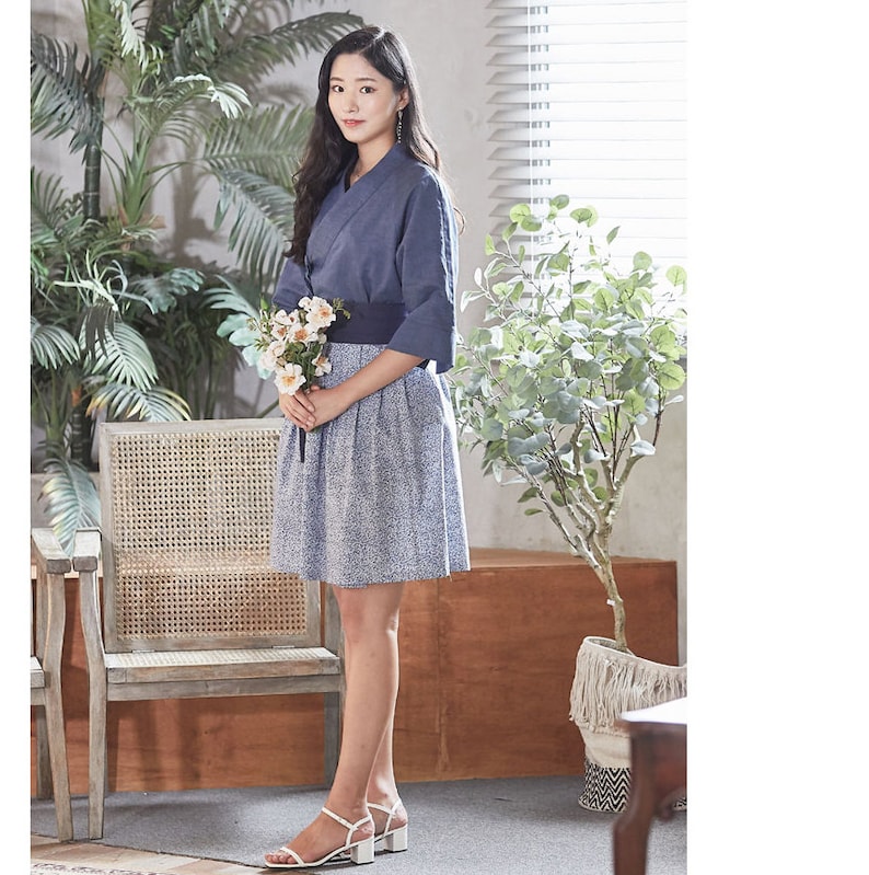May include: A woman wearing a navy blue wrap top with three-quarter sleeves, a matching wide belt, and a blue floral print skirt. She is holding a bouquet of white flowers and wearing white sandals with block heels. The outfit is suitable for a casual or semi-formal occasion.