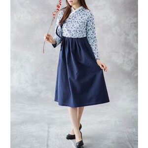 Op de afbeelding: Een jurk met lange mouwen met een lichtblauwe bloemenprint top en een donkerblauwe rok. De jurk heeft een traditioneel ontwerp met een hoge kraag en lange mouwen. Het model houdt een bloesemtak vast.