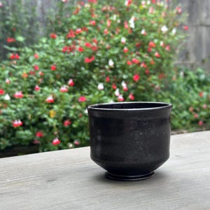 May include: A black ceramic teacup with a slightly flared rim and a rounded base. The cup is sitting on a wooden surface.