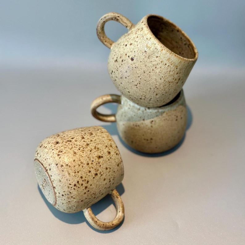 May include: Three speckled ceramic mugs with rounded bodies and curved handles are stacked and arranged on a light gray surface. The mugs have a neutral color palette with brown speckles and a light beige base. One mug is lying on its side.