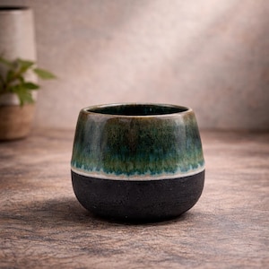 Pode incluir: Uma caneca de cerâmica artesanal com formato arredondado. A metade superior é esmaltada em tons de verde, passando para uma base preta. A caneca está sobre uma superfície de madeira, mostrando seu design único e contraste de cores.