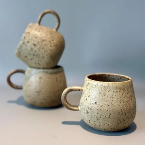 May include: A stack of three speckled ceramic mugs with rounded bodies and curved handles. The mugs have a neutral color palette with brown speckles, and the interior of the mugs is a darker shade. The mugs are arranged on a light gray surface.
