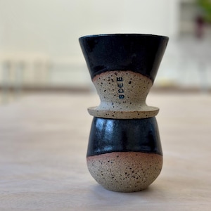 May include: A ceramic coffee pour-over dripper stacked on a matching mug. The set features a two-tone design with a glossy black glaze on the upper sections and a speckled, unglazed tan clay base. The word SDEB is stamped vertically on the dripper.