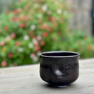 Puede incluir: Una taza de té de cerámica negra con un esmalte moteado. La taza está sobre una superficie de madera.