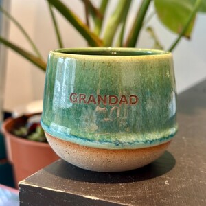 May include: A ceramic mug with a rounded shape, featuring a glossy green glaze on the upper portion and a textured, unglazed tan base. The word GRANDAD is embossed in brown capital letters on the front of the mug, centered within the green area.