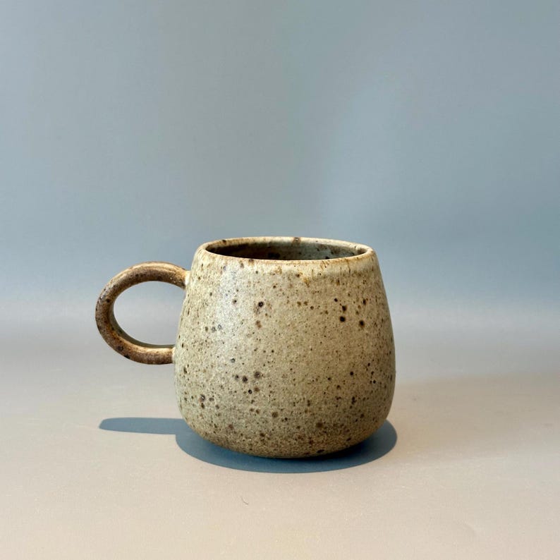 May include: A speckled ceramic mug with a rounded body and a sturdy, curved handle. The mug has a neutral color palette with brown speckles, and it sits on a light gray surface. The mug is designed for hot beverages.