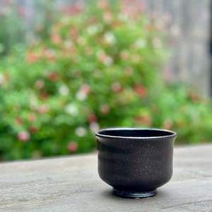 May include: A black ceramic cup with a speckled glaze sits on a wooden surface. The cup has a rounded shape and a slightly flared lip.