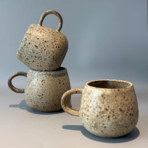 May include: Three speckled ceramic mugs with rounded bodies and curved handles are stacked and arranged on a gray surface. The mugs have a neutral color palette with brown and tan speckles, creating a rustic aesthetic. The mugs are ideal for coffee or tea.