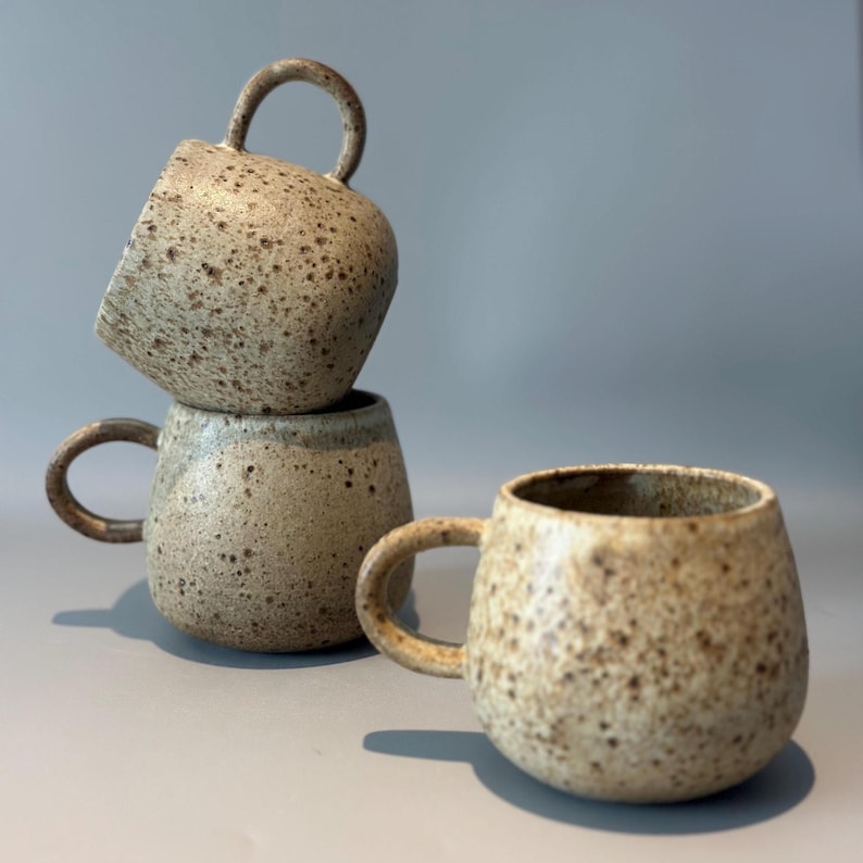 May include: Three speckled ceramic mugs with rounded bodies and curved handles are stacked and arranged on a gray surface. The mugs have a neutral color palette with brown and tan speckles, creating a rustic aesthetic. The mugs are ideal for coffee or tea.