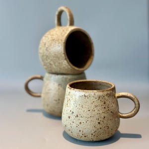 May include: Three speckled ceramic mugs in a neutral color palette. One mug has a rounded shape with a handle, while the other two are stacked, with one having a large opening and a handle. The mugs have a rustic, handmade appearance.