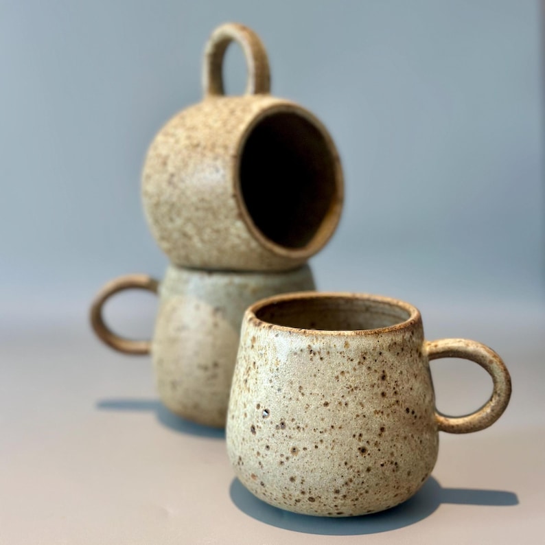 May include: Three speckled ceramic mugs in a neutral color palette. One mug has a rounded shape with a handle, while the other two are stacked, with one having a large opening and a handle. The mugs have a rustic, handmade appearance.