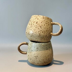 May include: Two handmade ceramic mugs stacked on top of each other. The mugs are beige with brown speckles and have rounded handles. The bottom mug has a light green band around the middle.