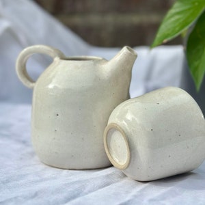 Könnte beinhalten: Ein weißes Keramik-Teekannen- und Tassenset. Die Teekanne hat einen runden Körper und einen Henkel. Die Tasse ist einfach und rund.