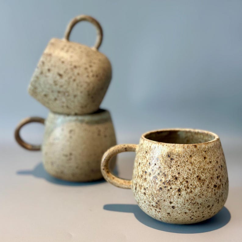 May include: Three speckled ceramic mugs in a stack. The mugs are a light beige color with brown speckles and rounded shapes. Each mug has a curved handle. The mugs are on a light gray surface.