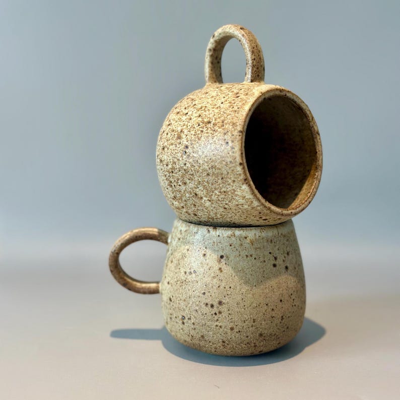 May include: Two speckled ceramic mugs stacked on top of each other. The top mug has a large handle and a circular opening. The bottom mug has a smaller handle. The mugs are beige with brown speckles.