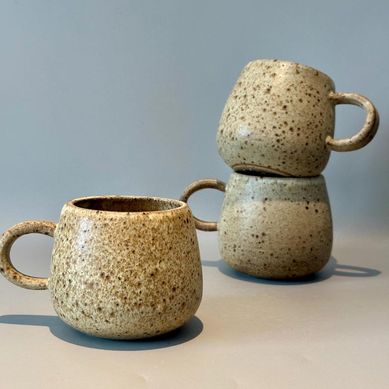 May include: Four speckled ceramic mugs with rounded bodies and curved handles. The mugs are stacked, with one on the bottom and two stacked on top. The mugs have a neutral color palette with brown and tan speckles.