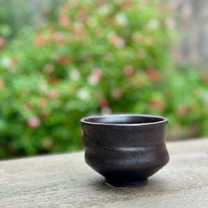 May include: A dark brown ceramic teacup with a rounded shape and a slightly flared rim. The cup is sitting on a wooden surface.