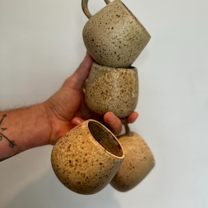 May include: Four speckled ceramic mugs in varying shades of tan and brown are stacked and held by a hand. Each mug has a rounded body and a small handle. The mugs have a rustic, handmade appearance.