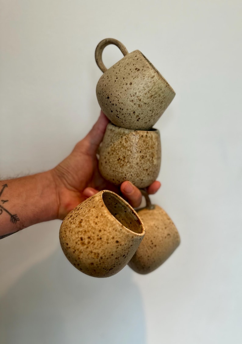 May include: Four speckled ceramic mugs in varying shades of tan and brown are stacked and held by a hand. Each mug has a rounded body and a small handle. The mugs have a rustic, handmade appearance.