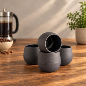 May include: A set of four rounded, matte black ceramic cups with a textured exterior and a smooth, dark interior. The cups are arranged in a cluster, with one cup positioned prominently on top of another, showcasing their simple, handleless design.