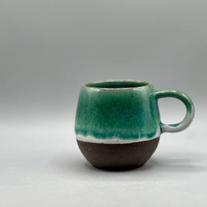 May include: A handcrafted ceramic mug with a rounded form and a handle. The upper section is glazed in a vivid teal hue, fading to a white band, and then a dark brown base. The mug is suitable for hot drinks.
