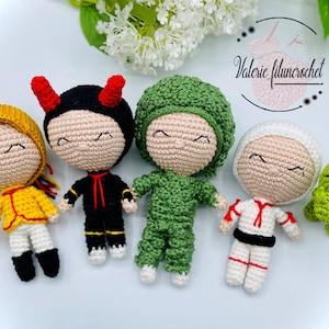 Puede incluir: Una colección de cinco muñecos de ganchillo, cada uno con un disfraz único. Los muñecos tienen los ojos cerrados y visten varios atuendos, incluyendo un atuendo amarillo con detalles rojos, un atuendo negro con cuernos rojos y un mono verde.