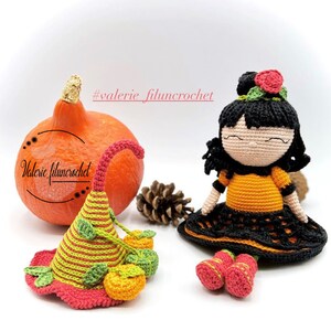 Puede incluir: Una muñeca de crochet que lleva un vestido negro y naranja con un sombrero de calabaza. La muñeca tiene el pelo negro y una cara sonriente. La muñeca está sentada junto a una pequeña calabaza.