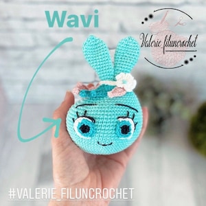 Peut inclure: Un jouet de lapin bleu en crochet avec un bandeau de fleurs blanches. Le lapin a un visage souriant avec des yeux noirs et des cils. Le jouet est tenu dans une main et le texte "Wavi" et "#VALERIE_FILUNCROCHET" sont visibles sur l'image.