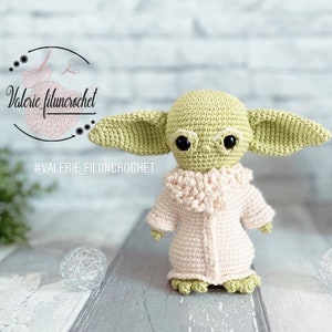 Peut inclure: Une poupée Yoda en crochet vert et rose portant un manteau en crochet blanc. La poupée est debout sur une surface en bois. #VALERIE_FILUNCROCHET