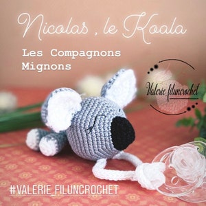 May include: A gray crocheted koala with white ears and a black nose, attached to a white crocheted pacifier clip. The koala is lying on a pink and white patterned surface. The text "Nicolas, le Koala Les Compagnons Mignons #VALERIE_FILUNCROCHET" is visible in the image.