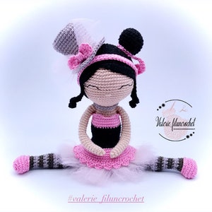 May include: A crocheted ballerina doll with black hair, a pink and black dress, and a gray top hat. The doll is sitting with her legs spread out and wearing a pink tutu.