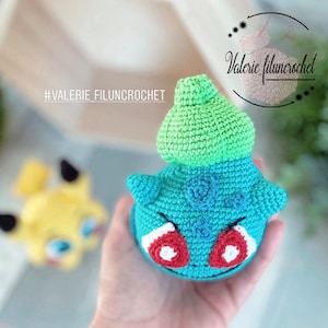 May include: A crocheted green and blue Pokemon character with red eyes. The character is being held in a hand. The hashtag #VALERIE_FILUNCROCHET is visible in the background.