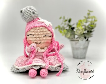 Pode incluir: Uma boneca de crochê feita com fio rosa e cinza, vestindo um vestido rosa e uma coroa cinza. A boneca tem um rosto sorridente e está sentada em uma superfície branca.