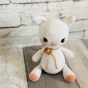 May include: White crocheted cat toy with a brown ribbon and a peach coloured nose. The cat is sitting on a wooden surface.