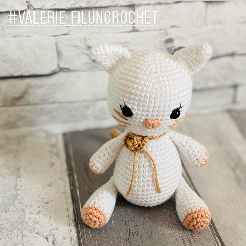 May include: White crocheted cat toy with a brown ribbon and a peach coloured nose. The cat is sitting on a wooden surface.