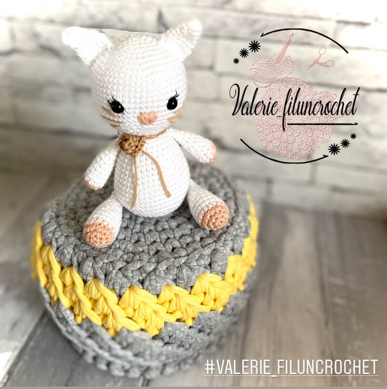 May include: White crocheted cat amigurumi toy with brown eyes and a brown yarn bow. The cat is sitting on a grey and yellow crocheted round cushion. #VALERIE_FILUNCROCHET