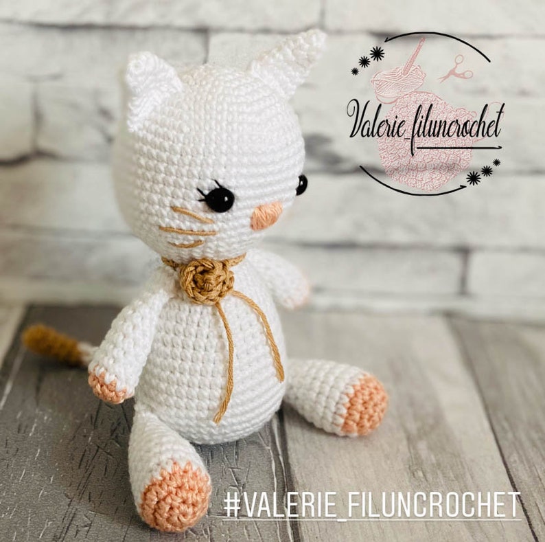 May include: White crocheted cat toy with a tan bow and peach-coloured paws. The cat is sitting on a brick surface with the text #VALERIE_FILUNCROCHET in the foreground.